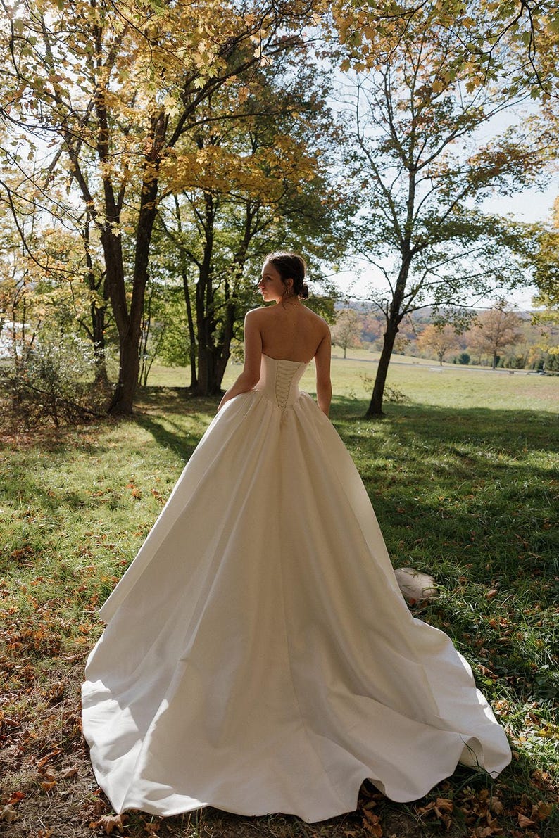 May include: A white strapless wedding gown with a fitted bodice and a full skirt. The gown has a lace-up back and is made of a smooth, satin fabric.