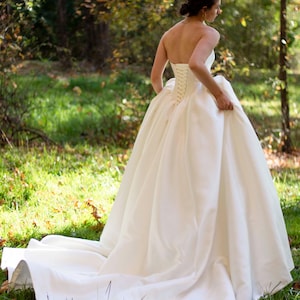 May include: A white strapless wedding dress with a fitted bodice and a full, flowing skirt. The dress has a lace-up back and is made of a smooth, satin fabric.