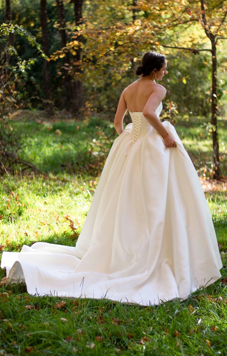 May include: A white strapless wedding dress with a fitted bodice and a full, flowing skirt. The dress has a lace-up back and is made of a smooth, satin fabric.