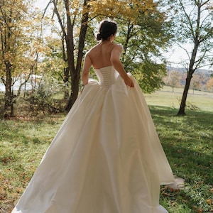 May include: A white strapless wedding dress with a fitted bodice and a flowing skirt. The dress is made of a smooth, silky fabric and has a simple, elegant design. The dress is being worn by a person in a grassy outdoor setting.