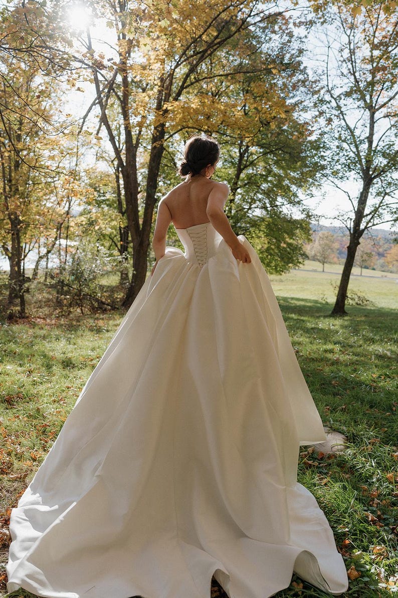 May include: A white strapless wedding dress with a fitted bodice and a flowing skirt. The dress is made of a smooth, silky fabric and has a simple, elegant design. The dress is being worn by a person in a grassy outdoor setting.