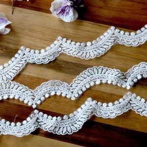 May include: White lace trim with a scalloped edge and a floral design. The trim is arranged in wavy lines on a wooden surface, with purple and white flowers in the background. This delicate lace is suitable for sewing and craft projects.