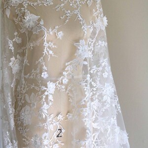 May include: White floral lace fabric with delicate embroidery and a sheer texture. The lace features a floral pattern with small and large flowers, creating a romantic and elegant look.
