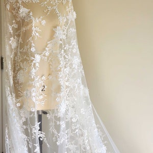 May include: White floral lace fabric with a delicate pattern of flowers and leaves. The lace is sheer and has a soft, flowing texture.