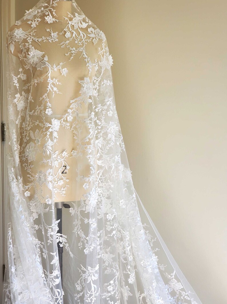 May include: White floral lace fabric with a delicate pattern of flowers and leaves. The lace is sheer and has a soft, flowing texture.