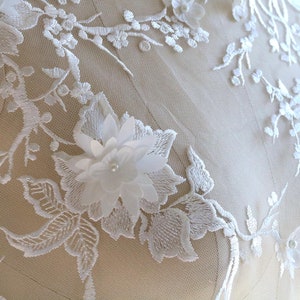 May include: White lace fabric with delicate floral embroidery and a large 3D flower applique. The fabric is sheer and has a soft, romantic feel.