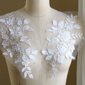 May include: Two white floral lace appliques with beaded details. The appliques are on a white mesh background.