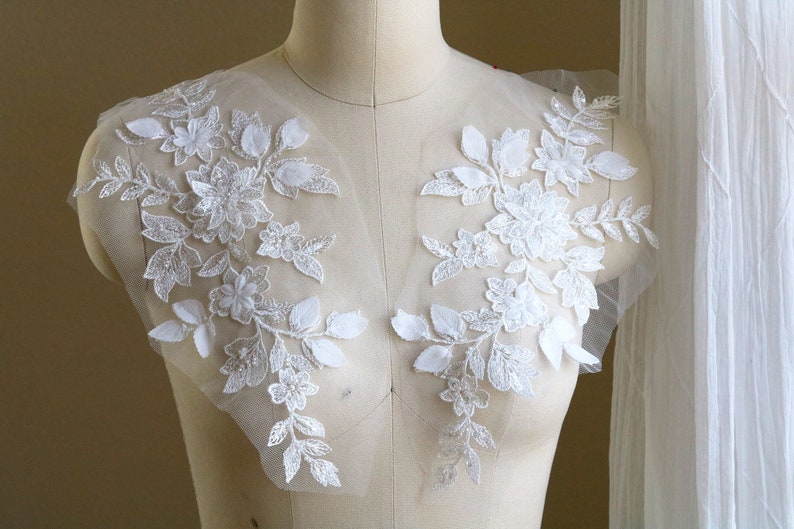 May include: Two white floral lace appliques with beaded details. The appliques are on a white mesh background.