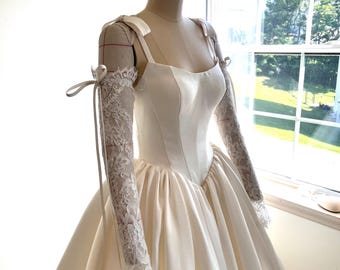 May include: Ivory wedding gown on a mannequin. The dress features a fitted bodice, a full skirt, and delicate lace sleeves tied with ribbons. The gown is displayed near a window, showcasing its elegant design.