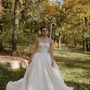 May include: A white strapless wedding dress with a full skirt and a long train. The dress is made of a smooth, satin fabric. The bride is standing in a grassy area with trees in the background.