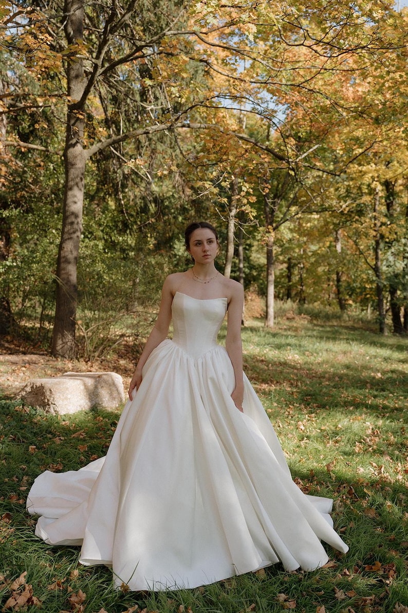 May include: A white strapless wedding dress with a full skirt and a long train. The dress is made of a smooth, satin fabric. The bride is standing in a grassy area with trees in the background.