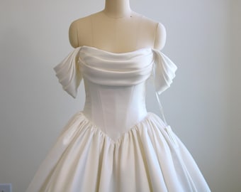 May include: A white wedding dress on a mannequin. The off-the-shoulder bodice has draped fabric over the shoulders. The skirt is full and gathered, creating a voluminous silhouette. The dress is made of a smooth, satin-like material.
