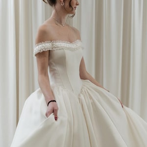 May include: Off-the-shoulder ivory wedding gown with a fitted bodice and full skirt. The dress features a delicate lace trim along the neckline and a classic, elegant silhouette. The bride's hair is styled in an updo.