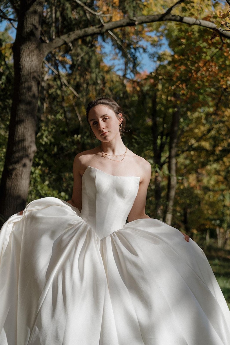 May include: A woman wearing a white strapless wedding dress with a sweetheart neckline. The dress has a full skirt and is made of a smooth, satin fabric. The woman is standing in a forest setting with trees and leaves in the background.