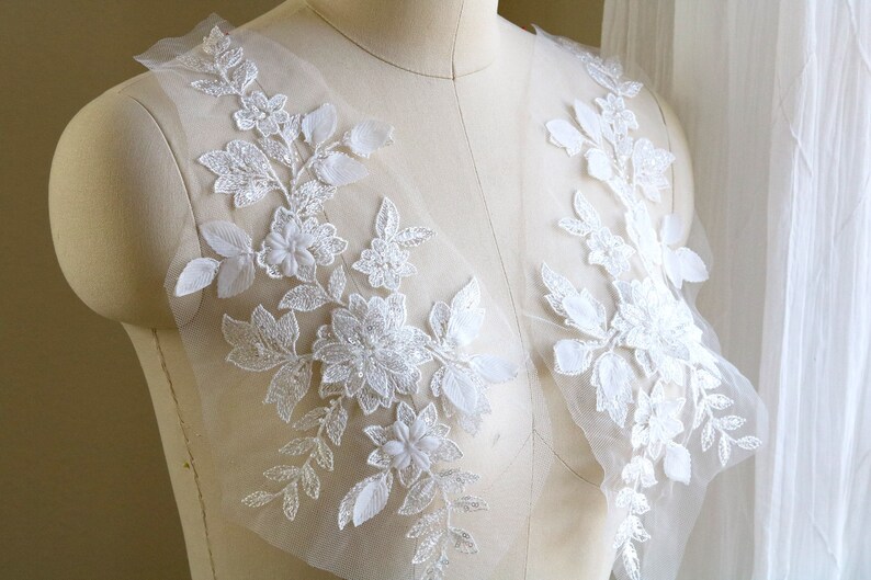 May include: Two white floral lace appliques with beaded details on a sheer white fabric background.