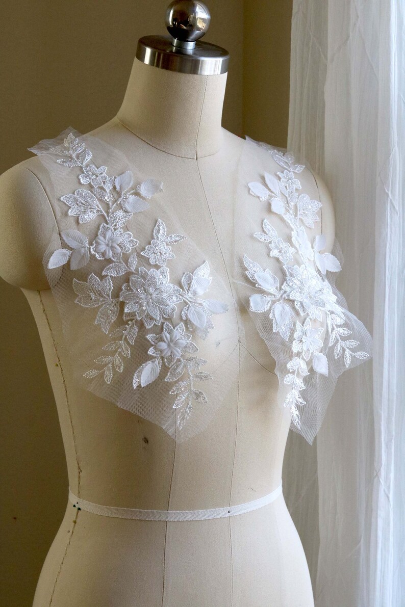 May include: Two white floral lace appliques on a white dress form. The appliques have a delicate floral design with small white flowers and leaves. The appliques are made of a sheer fabric with a subtle shimmer.