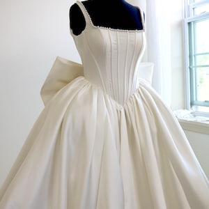May include: Ivory-colored wedding gown with a structured bodice and a full skirt. The dress features a square neckline trimmed with small pearls and a large bow detail at the back. The gown is displayed on a black mannequin.