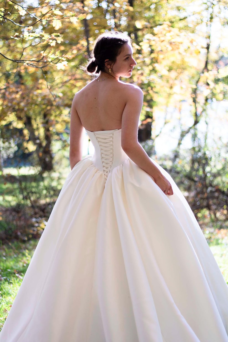 May include: A white strapless wedding dress with a lace-up back detail. The dress has a full skirt and is made of a smooth, satin fabric.