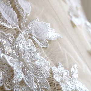 May include: Close-up of white lace fabric with delicate floral patterns and sequins. The fabric has a soft, sheer texture and a subtle shimmer.