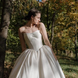 May include: A white strapless wedding dress with a fitted bodice and a full skirt. The dress is made of a smooth, flowing fabric and has a simple, elegant design.