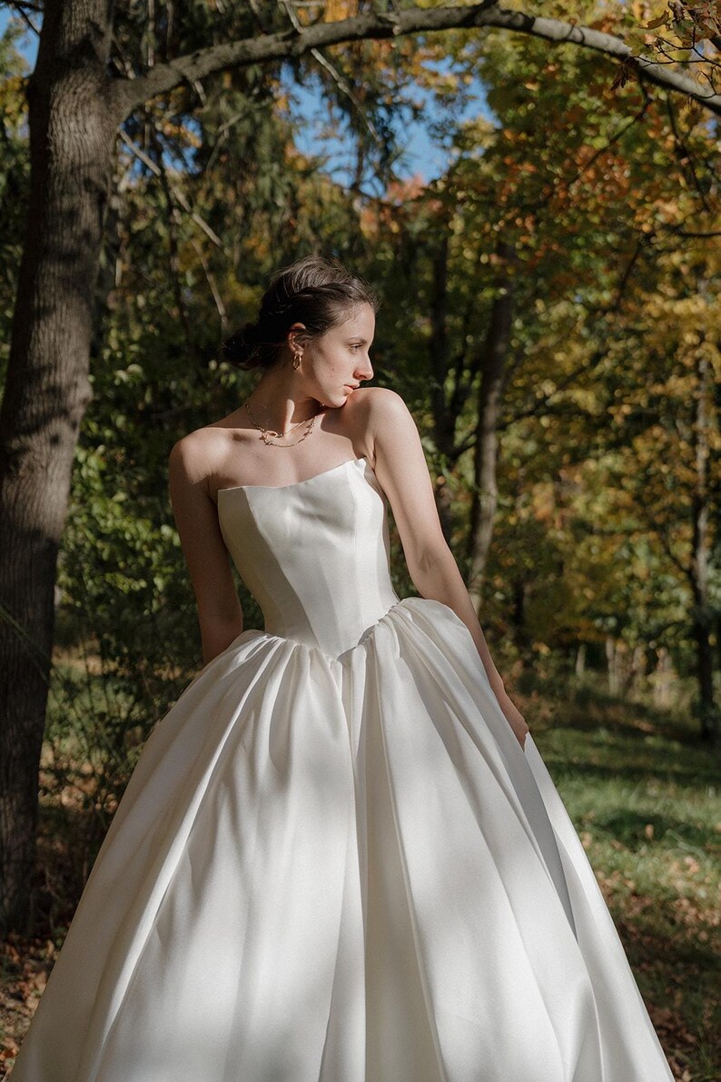 May include: A white strapless wedding dress with a fitted bodice and a full skirt. The dress is made of a smooth, flowing fabric and has a simple, elegant design.