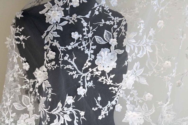 May include: White floral lace fabric with delicate 3D flowers and intricate designs. The lace is soft and sheer, perfect for adding a touch of elegance to any project.