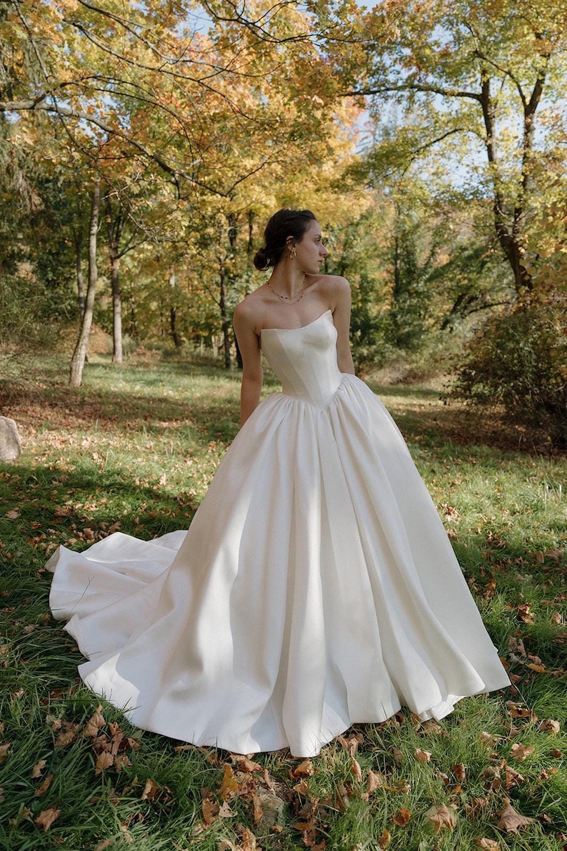 May include: A white strapless wedding dress with a fitted bodice and a full skirt. The dress is photographed in a forest setting with fall foliage in the background.