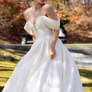 May include: A bride wearing a white off-the-shoulder wedding dress with a lace bodice and a flowing skirt. The dress is photographed outdoors in a natural setting with autumn foliage in the background.
