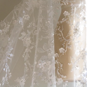 May include: White floral lace fabric with delicate 3D flowers and pearl accents. The lace is draped over a dress form.