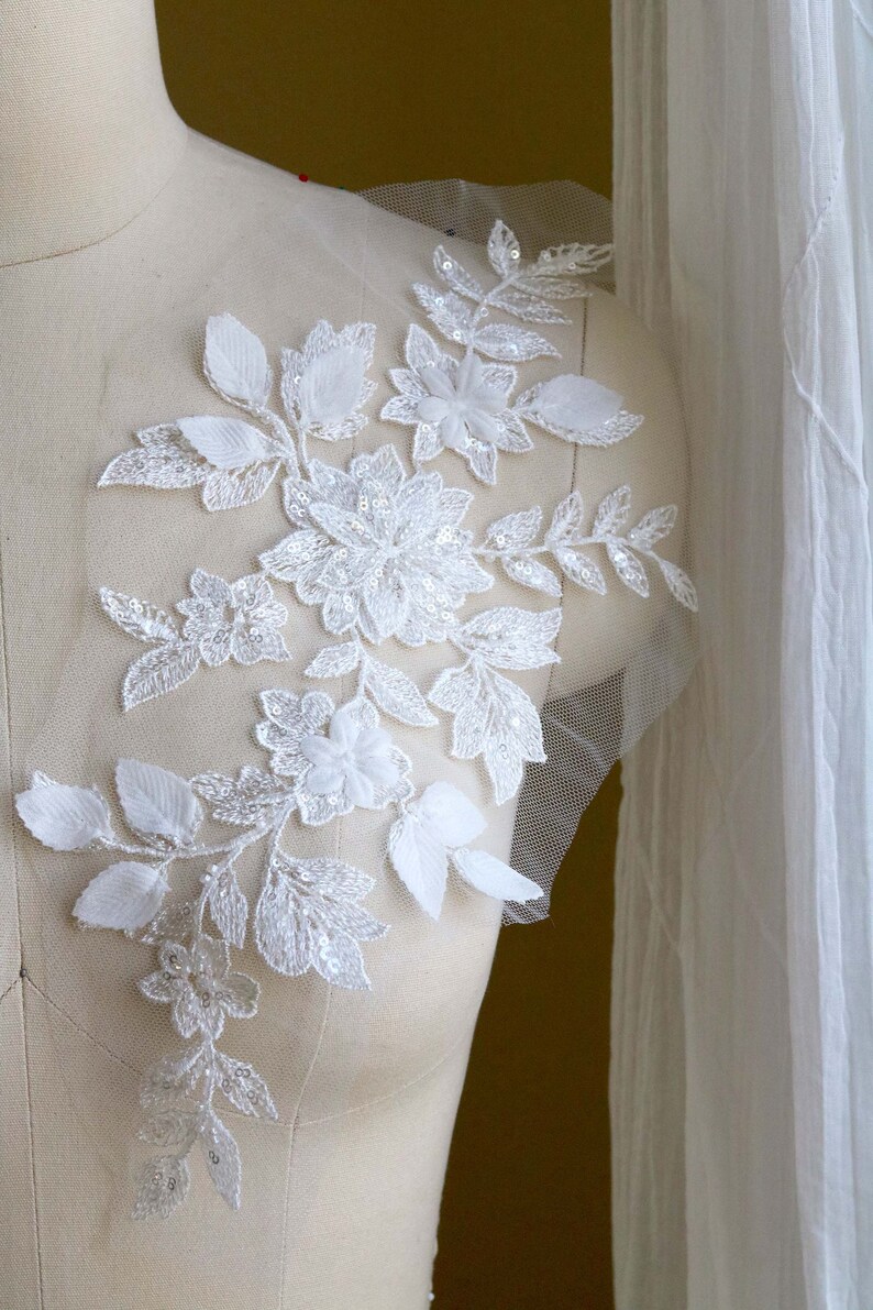 May include: White floral lace applique with sequins and beads. The applique is on a white mesh fabric and has a delicate design with leaves and flowers.