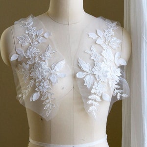 May include: Two white floral lace appliques with sequins on a white dress form.