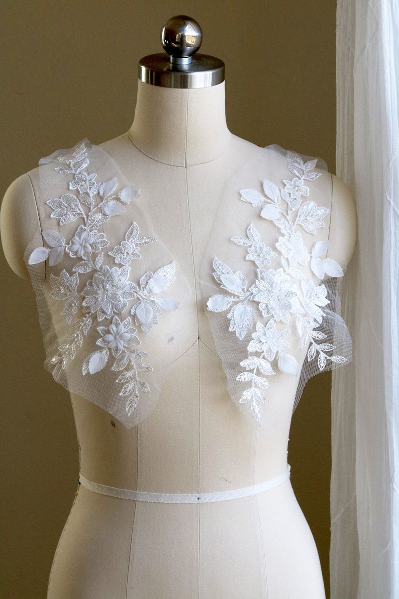 May include: Two white floral lace appliques with sequins on a white dress form.