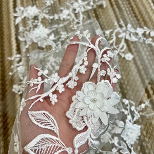 May include: White floral lace fabric with a delicate floral pattern and 3D flowers. The lace is soft and sheer, and the flowers are made of a white fabric with a pearl center.