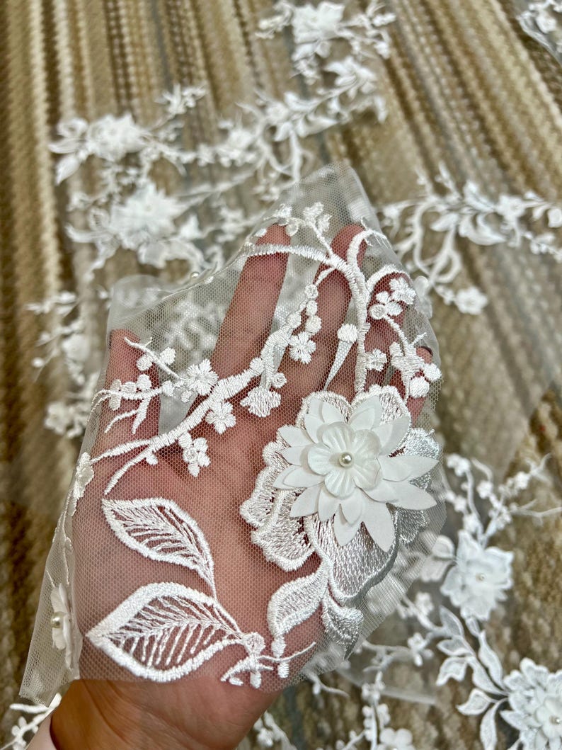 May include: White floral lace fabric with a delicate floral pattern and 3D flowers. The lace is soft and sheer, and the flowers are made of a white fabric with a pearl center.