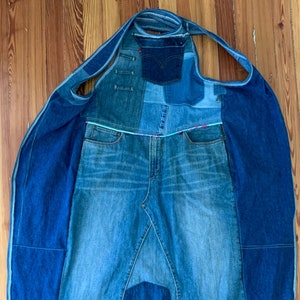 May include: A denim tote bag made from repurposed jeans. The bag features a patchwork design with various shades of blue denim. It has a large main compartment, a small pocket, and shoulder straps. The bag is approximately 76 cm long.
