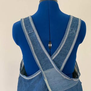 May include: A blue denim apron featuring wide, crisscross shoulder straps that form an X-shape on the back. The garment is constructed from medium-wash denim fabric with visible light-coloured stitching along the edges of the straps.