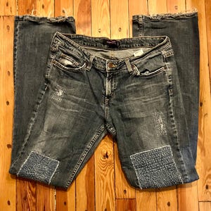 May include: Medium-wash blue denim jeans featuring a low-rise waist, a button and zipper fly, and five pockets. The jeans have distressed areas with frayed edges, and both legs feature rectangular patches with white stitched grid patterns.