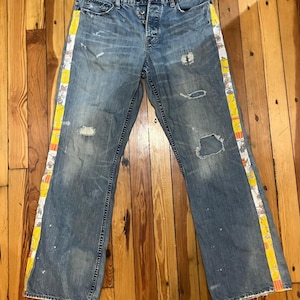 May include: American Eagle blue denim jeans featuring a button fly, distressed areas with rips, and vertical side panels decorated with a yellow and white floral pattern. The jeans have a faded wash, white paint splatters, and a classic five-pocket design.