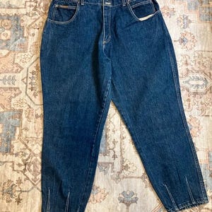 Può includere: Jeans in denim blu scuro caratterizzati da un design a vita alta con fascia elastica in vita, chiusura frontale con bottone e cerniera, due tasche laterali e dettagli con cuciture verticali sul fondo di ogni gamba.