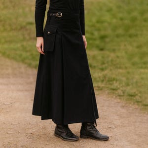 May include: A long, black skirt with a buckle belt and a small pocket. The skirt has a flowing, A-line shape and is paired with black boots. The outfit is suitable for various occasions.