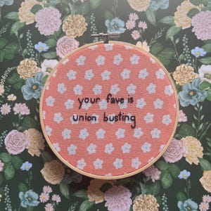 May include: A circular wooden embroidery hoop framing a coral-colored fabric with a repeating pattern of small white flowers. The text "your fave is union busting" is embroidered in black thread across the center of the fabric.