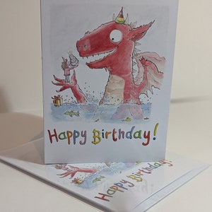 Op de afbeelding: Verjaardagskaart met een cartoon illustratie van een rode draak met een feesthoedje en een persoon die een taart aanbiedt. De woorden "Happy Birthday!" zijn in kleurrijke letters geschreven. Een andere kaart is zichtbaar.