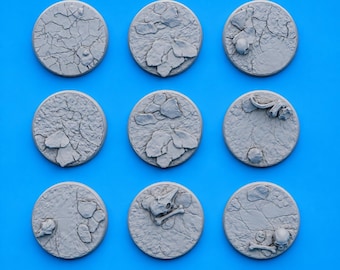 Op de afbeelding: Negen grijze, cirkelvormige miniatuurbasen met een getextureerd oppervlak. Sommige basen hebben een patroon van gebarsten aarde, terwijl andere rotsformaties, schedels en botten bevatten. De basen zijn gerangschikt op een helderblauwe achtergrond.