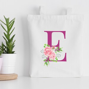 K&ouml;nnte beinhalten: Wei&szlig;e Einkaufstasche mit einem rosafarbenen Blumendesign und dem Buchstaben "E" in Magenta.