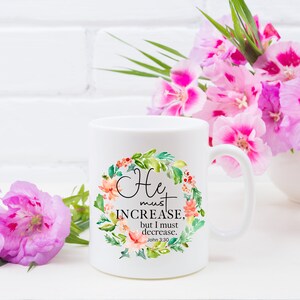 Pu&ograve; includere: Tazza in ceramica bianca con un design a corona di fiori e il testo "He must increase, but I must decrease. John 3:30".