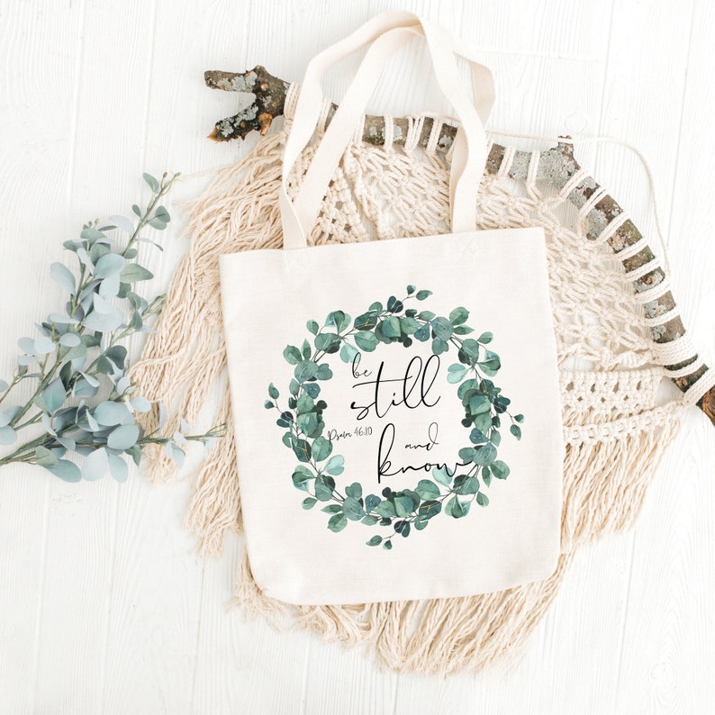Puede incluir: Una bolsa de lona blanca con un dise&ntilde;o de corona de eucalipto verde. La corona tiene las palabras "be still and know" en texto negro con la referencia b&iacute;blica "Salmo 46:10" debajo.