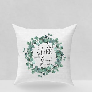 Puede incluir: Coj&iacute;n blanco con un dise&ntilde;o de corona de eucalipto verde. La corona tiene las palabras "be still and know" en texto negro con la referencia b&iacute;blica "Salmo 46:10" debajo.