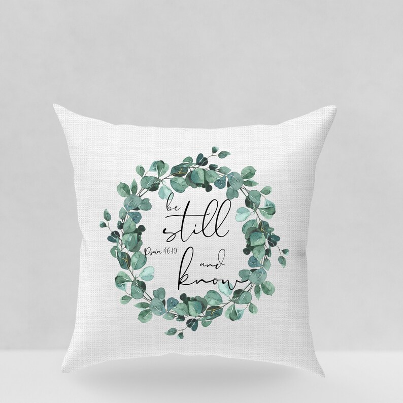 Puede incluir: Coj&iacute;n blanco con un dise&ntilde;o de corona de eucalipto verde. La corona tiene las palabras "be still and know" en texto negro con la referencia b&iacute;blica "Salmo 46:10" debajo.