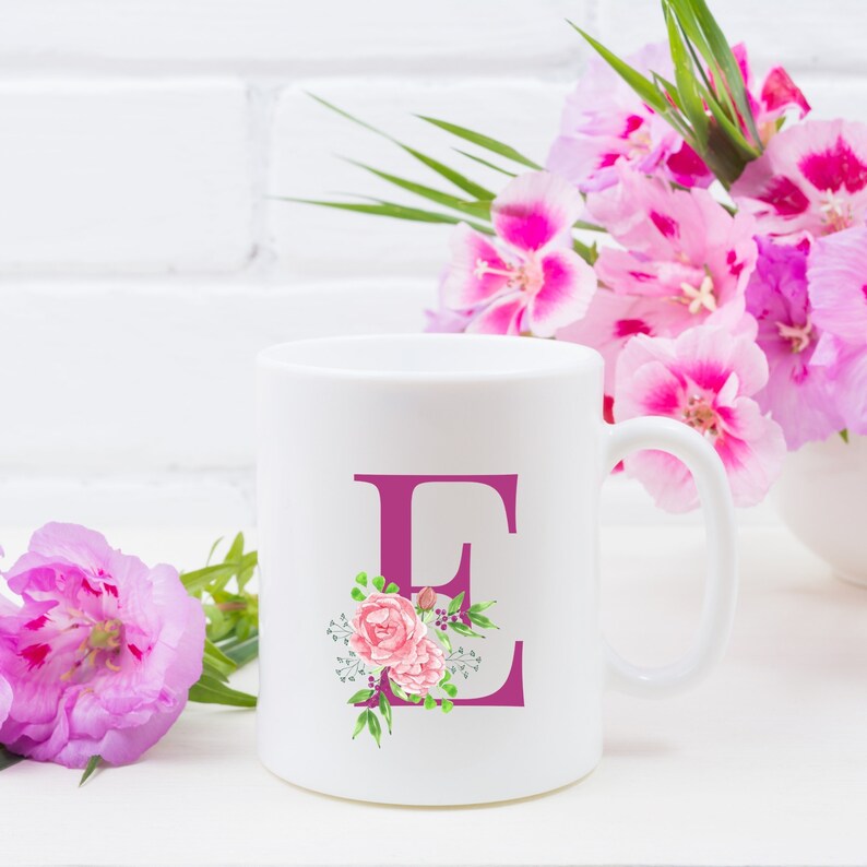 K&ouml;nnte beinhalten: Wei&szlig;e Keramiktasse mit einem rosafarbenen Blumendesign und dem Buchstaben "E" in einem dunkleren Rosa. Die Tasse steht auf einer wei&szlig;en Oberfl&auml;che mit rosafarbenen Blumen im Hintergrund.
