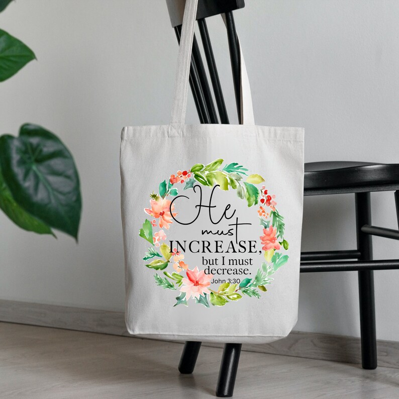 Pu&ograve; includere: Una borsa tote in tela bianca con un design a corona di fiori e il testo "He must increase, but I must decrease. John 3:30".